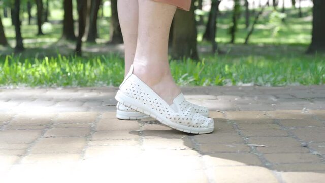 Camera follows senior female Caucasian feet walking along alley in sunny park. Unrecognizable woman stopping because of ankle pain. Medical problems and aging concept.