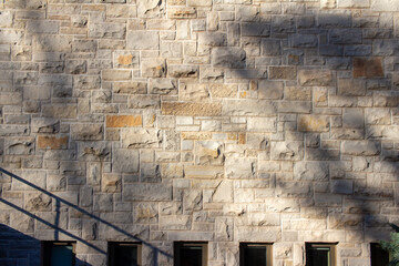 Full frame abstract background of an attractive tan brown natural limestone block wall in ashlar pattern, with rugged texture stone blocks in full sunlight with tree shadows and partial view of window