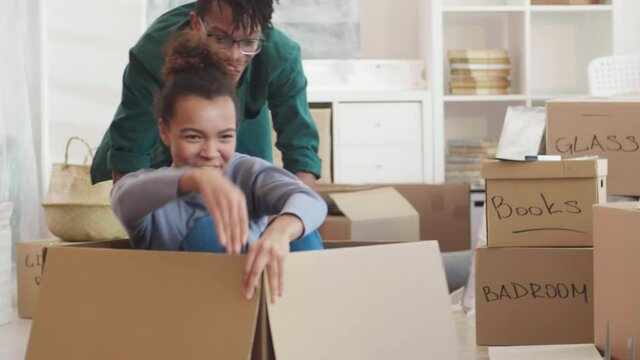 Medium shot of young mixed-race woman sitting in cardboard box and having fun with her handsome boyfriend in moving day