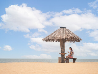 Asian woman sitting under straw umbrella on seashore. female using mobile smart phone on beach. copy space provided.