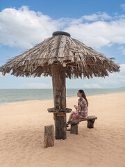Asian woman sitting under straw umbrella on tropical beach. female looking to sea view.