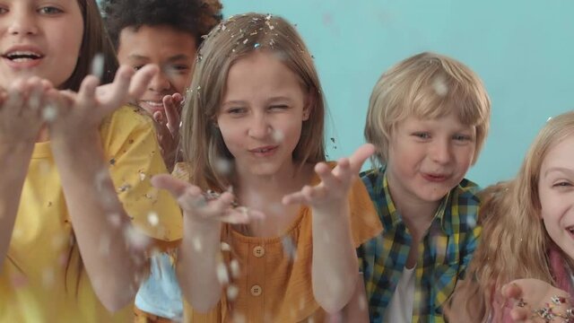 Medium close-up POV of five multiethnic school kids holding handful of sparkles, breathing in deeply and blowing it off on camera. Girls and boys smiling and enjoying time standing on blue background