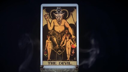A tarot Devil card sits on a brass stand as smoke rises around it. The mystical satanic figure is displayed on a black background. Satan sits on his evil smokey throne.