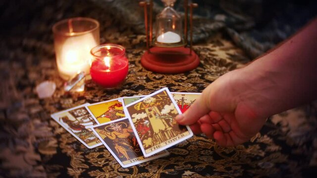 A mystical fortune teller lays out a tarot card spread. The devil, tower, and wheel of fortune cards are displayed on a mysterious table adorned with candles, and hourglass and crystals.