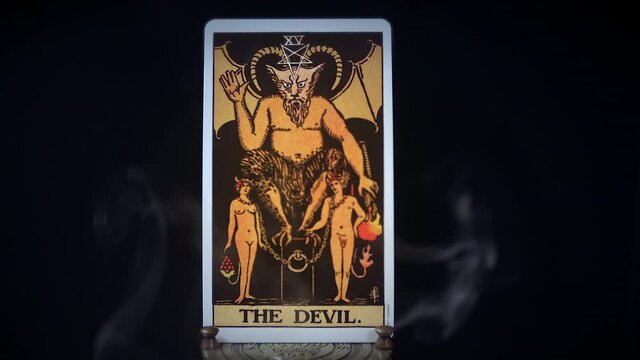 A tarot Devil card sits on a brass stand as smoke rises around it. The mystical satanic figure is displayed on a black background. Satan sits on his evil smokey throne.