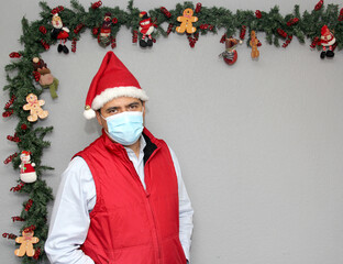 
latin man with protection mask, red vest and santa claus hat in christmas decoration, new normal...
