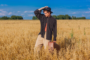 Naklejka na ściany i meble Tall handsome man with black hat and brown suitcase standing at golden oat field.