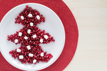 Christmas tree made of pomegranate seeds on plate on table with small marshmallow as decorations. Healthy snack and creative christmas food concept. 