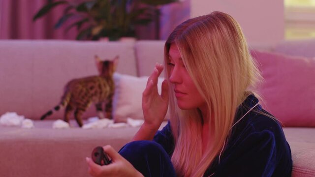 Miserable unhappy stressful blonde woman starts to cry while watching drama movie on TV. Her adorable bengal cat playing funny at background. Emotions. At home.