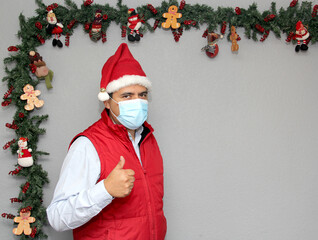
latin man with protection mask, red vest and santa claus hat in christmas decoration, new normal...