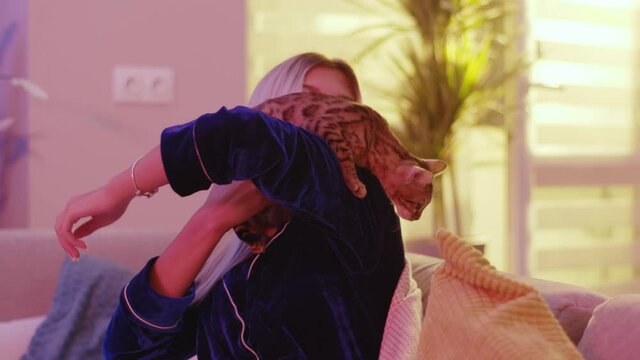 Blonde Caucasian Woman In Blue Pajama Playing With Lovely Kitten At Home. Funny Curious Playful Bengal Cat Trying To Escape From Mommy. Pets. Cute Animals.