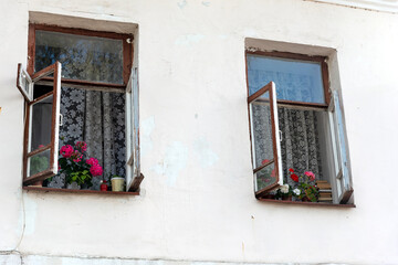 Fototapeta na wymiar Rustic house with white washed walls and open windows. Blooming potted flowers on sills. Summer cottage or old styled vintage dwelling in countryside. Retro apartment exterior, facade of country flat