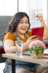 Obraz na płótnie Canvas portrait of fat asian woman wearing casual cloth sitting on the floor in a house holding a tablet and hand up looking at a camera with smiling face. Selective focus on a happy lady face