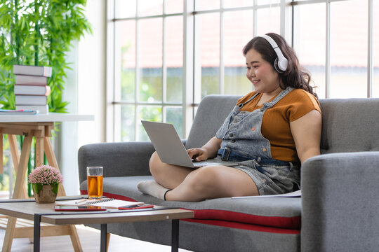  Fat Female Wearing Casual Cloth With Headphones Smiling And Sitting On A Couch Looking At Laptop Computer On Her Lap During Doing Online Communication In A House. Study And Work From Home Concept