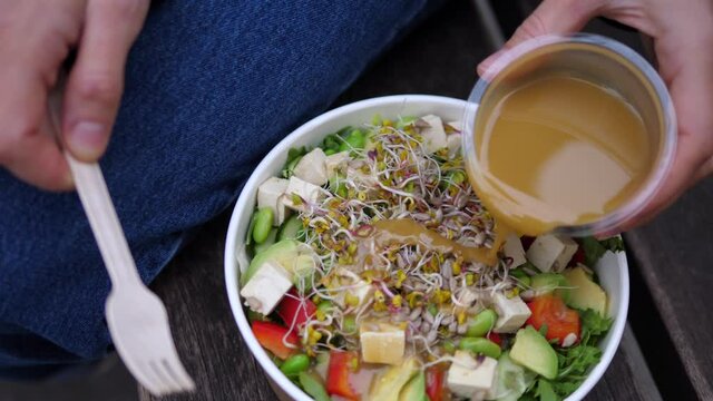 Top view of hand dressing raw vegan salad with tahini oil in single use container. Healthy snack for lunch break outdoors 