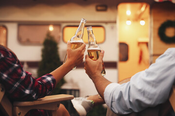 Young couple toasting with bottles of beer near trailer, closeup. Camping season
