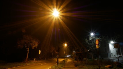 Einsame Dorfstraße in der Nacht im Nebel mit Strassenlaternen mit Blendenstern, die ein Gefühl...