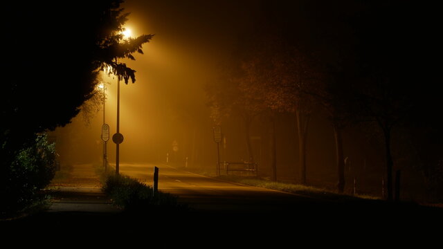 Einsame Landstra&szlig;e in der Nacht im dichten Nebel mit Strassenlaternen, die ein fahles Licht erzeugen und ein Gef&uuml;hl von Dystopie, Grusel, Einsamkeit einer Pandemie wie Corona
