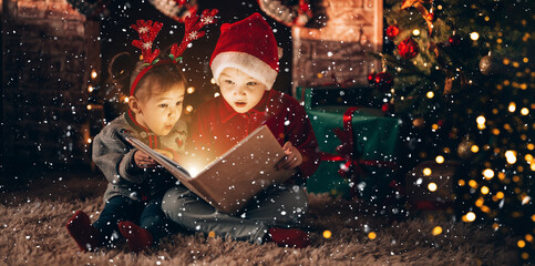 Brother and sister are sitting and reading a book. Christmas mood. Children in New Year's costumes against the background of a Christmas tree and a fireplace
