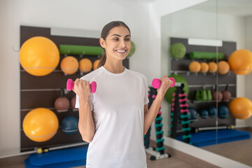 Smiling dark-haired female lifting dumbbells in the gym