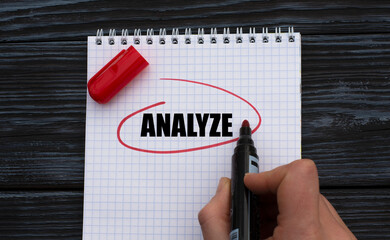 A woman's hand writes on a white sheet the word ANALYZE on a black wooden background