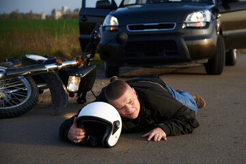 A motorcyclist lies on the asphalt near a motorcycle and car.