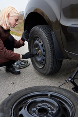 A young blonde woman removes the wheel with a key near her car with a flat tire.