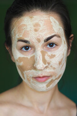 young brunette woman with a clay homemade mask on her face looks into the frame