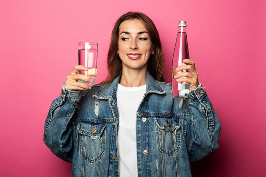 Friendly young woman holding a glass of water and a bottle of water on pink background.