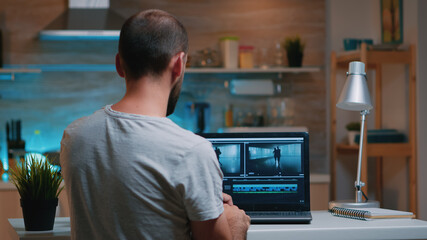 Colorist working remotely overtime from home using editing software sitting in front of laptop late at night. Videographer processing audio film montage on professional in modern kitchen in midnight