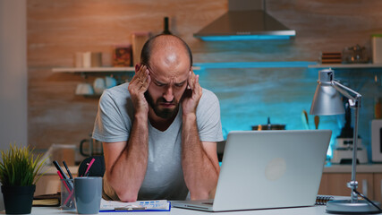 Freelancer suffering from headache touching temple after hours of work on laptop at home in modern...