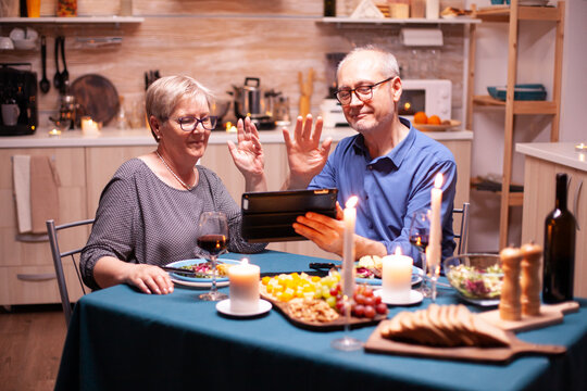 Mature man and woman waving at tablet pc during video conference in kitchen celebrating relationship. Couple sitting at the table, browsing, talking, using internet, celebrating their anniversary in