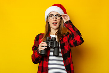 Young woman with a surprised face and in a Santa hat and a red shirt in a cage holds binoculars in her hands on a yellow background. Banner
