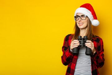 Young woman with a smile in a Santa hat and a red shirt in a cage looks to the side and holds binoculars in her hands on a yellow background