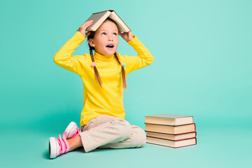 Full body photo of amazed little girl hide book on head look empty space wear yellow turtleneck...