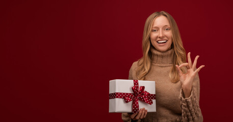 Charming young blonde woman shows the OK sign and holds gift in her hand. Studio shot red background. Mock up copy space