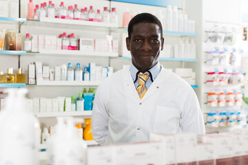 smiling male pharmacist standing on background with shelves of medicines in pharmacy