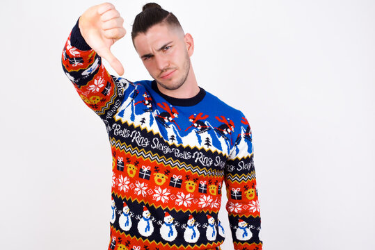 Discontent Young handsome Caucasian man wearing Christmas sweater against white wall shows disapproval sign, keeps thumb down, expresses dislike, frowns face in discontent. Negative feelings.