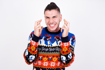 Young handsome Caucasian man wearing Christmas sweater against white wall holding fingers crossed...