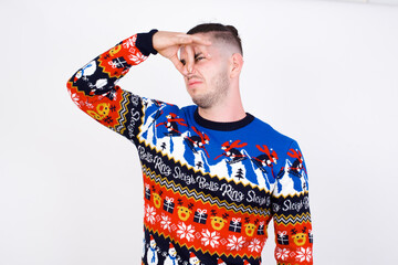 Displeased Young handsome Caucasian man wearing Christmas sweater against white wall plugs nose as...