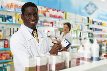 smiling confident adult African American pharmacist taking inventory of medicines in pharmacy