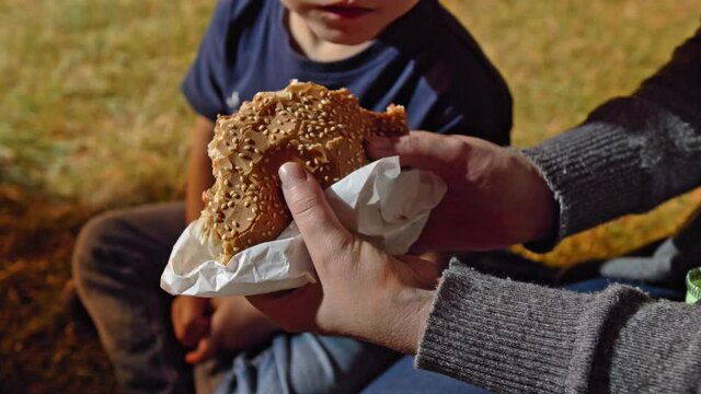 Woman holding  hamburger in hands. Nearby, the child is chewing food. They check what's inside. Mom and boy sitting on the ground outside.