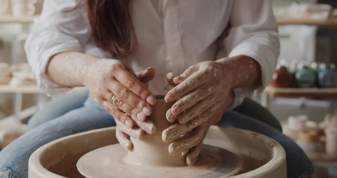 Loving couple enjoying romantic date in pottery workshop. Lovers working together on a pottery wheel in front of shelves with clay dishes. Close-up shot of hands.