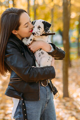 Naklejka na ściany i meble Woman holds and kisses puppy of dalmatian dog in autumn park.