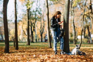 Woman looks in smartphone next to puppy of dalmatian dog in autumn park.