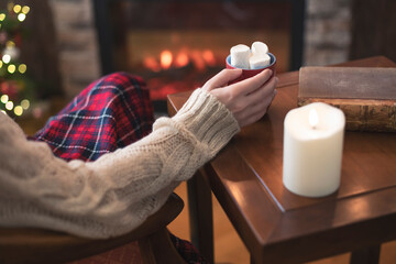 Woman hand with old retro book and cup of hot cocoa and marshmallow on wooden table near christmas tree and fireplace. Toned