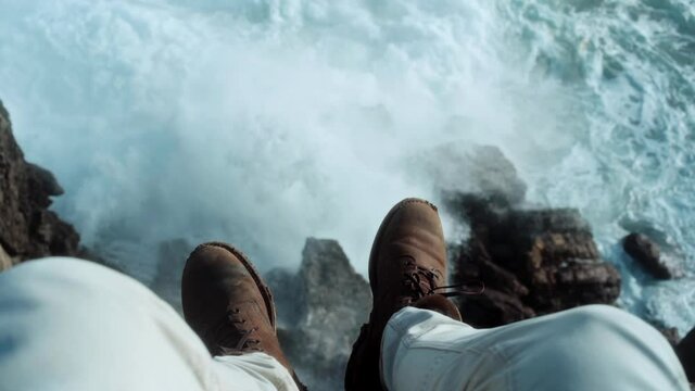 Cinematic shot of man in leather suede shoes sit on edge of cliff look down at waves and rocks. Adventure wanderlust concept, millennial travel ideas. Moody atmosphere of alone time in nature