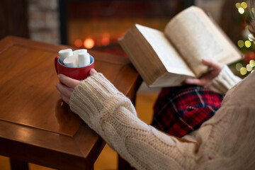 Fototapeta na wymiar Woman hand with old retro book and cup of hot cocoa and marshmallow on wooden table near christmas tree and fireplace.