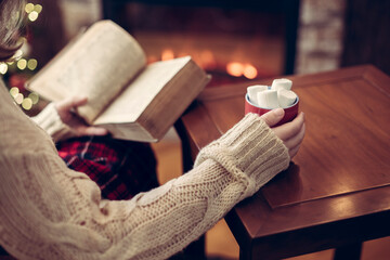Fototapeta na wymiar Woman hand with old retro book and cup of hot cocoa and marshmallow on wooden table near christmas tree and fireplace. Toned
