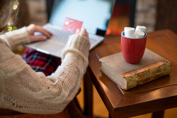 Woman  holding credit card using laptop for making order sitting at table with cup of hot cocoa and marshmallow at christmas fireplace with decoration of light bulbs. Close up.